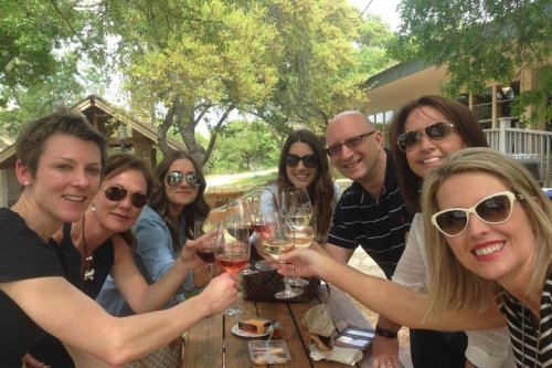 A group of seven friends cheerfully toasting with wine glasses outdoors, enjoying a sunny day together.