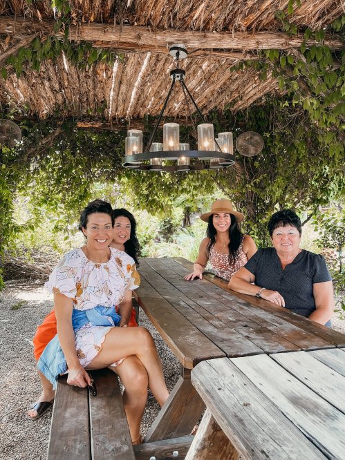 Four women sit at a wooden table under a leafy canopy, smiling and enjoying a sunny day outdoors.