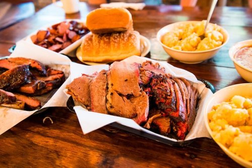 A spread of barbecue meats, including brisket and sausage, with sides of macaroni and cheese and bread rolls on a wooden table.