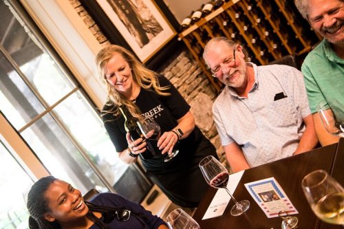 A group enjoying wine tasting at a winery, with a server pouring wine for guests at a table.