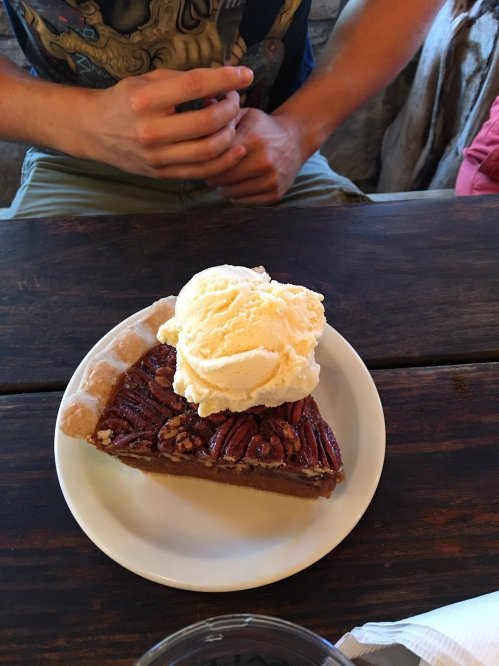 A slice of pecan pie topped with a scoop of vanilla ice cream on a white plate, resting on a wooden table.