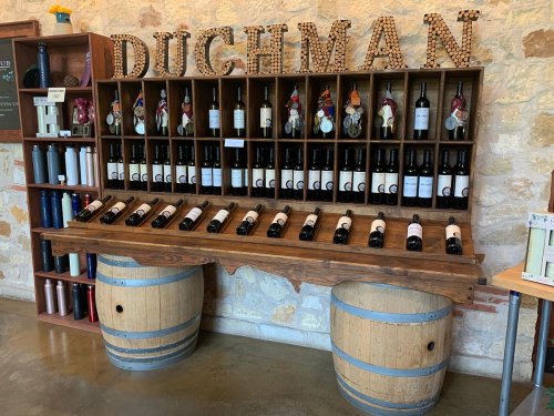 A wooden wine display with bottles arranged on a shelf, featuring the name "DUCHMAN" above, set against a stone wall.