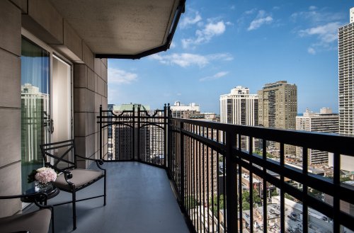 A balcony view overlooking a city skyline with tall buildings and a clear blue sky. A chair and small table are visible.