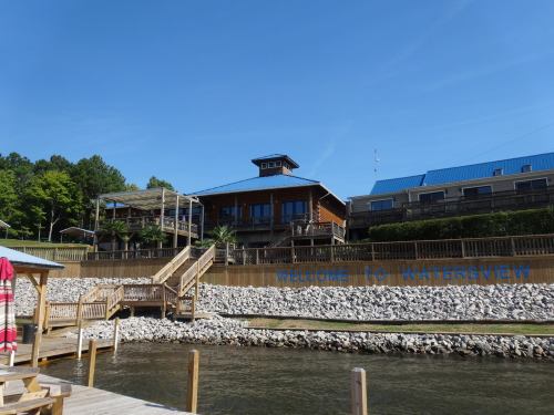 A wooden building with a deck overlooks a calm waterway, surrounded by trees and blue skies. "Welcome to Watersview" sign visible.