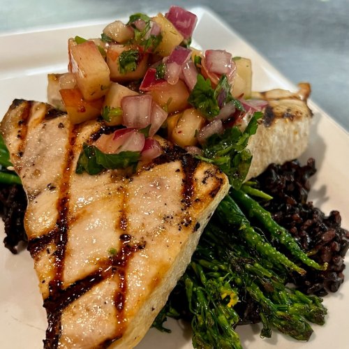 Grilled fish fillet topped with fruit salsa, served with black rice and sautéed greens on a white plate.