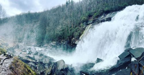A scenic waterfall cascades over rocks, surrounded by a misty forest landscape under a cloudy sky.
