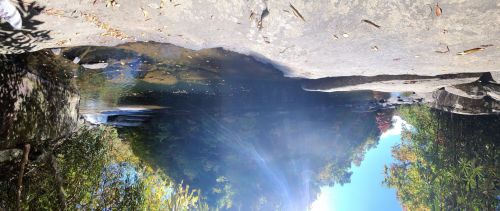 A serene natural scene featuring a calm water pool surrounded by rocks and lush greenery under a clear blue sky.