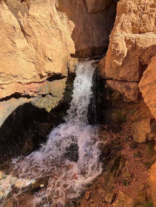 A small waterfall cascades over rocky terrain, surrounded by rugged, golden-brown cliffs.