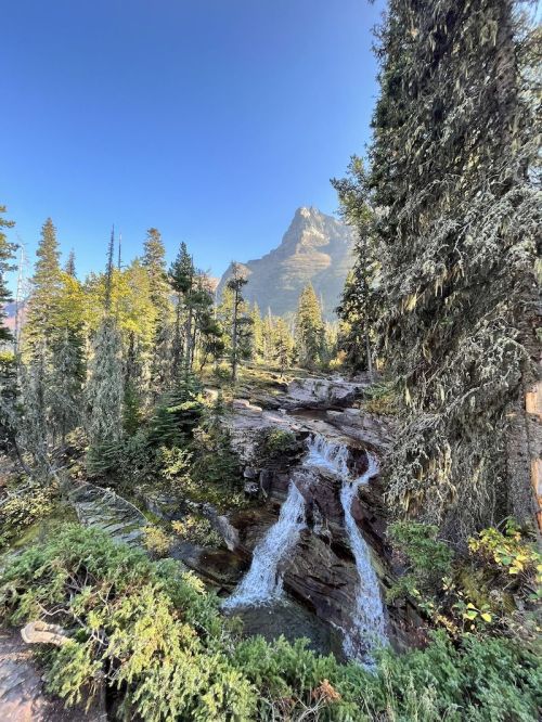 A serene landscape featuring a waterfall surrounded by lush trees and a mountain in the background under a clear blue sky.