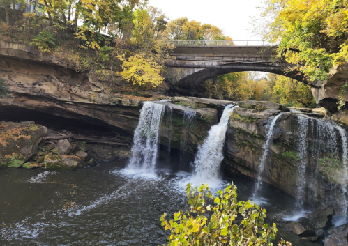 A scenic view of waterfalls cascading over rocks, with a bridge above and autumn foliage surrounding the area.