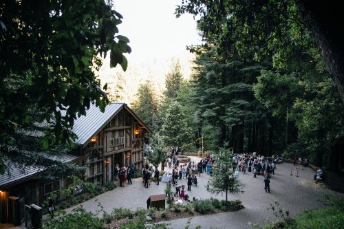 A scenic outdoor gathering at a rustic venue surrounded by tall trees, with guests mingling and enjoying the atmosphere.