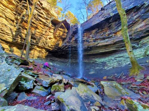 A waterfall cascades down rocky cliffs surrounded by trees, with a person exploring the area below.