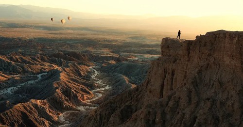 A person stands on a rocky cliff overlooking a vast desert landscape with hot air balloons in the distance at sunset.