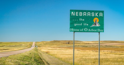 State welcome sign reads "Nebraska...the good life, Home of Arbor Day" against a wide-open blue sky.