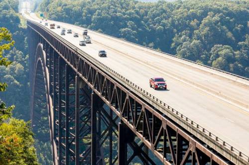A red truck drives on a high bridge with cars, surrounded by lush green trees and a clear blue sky.