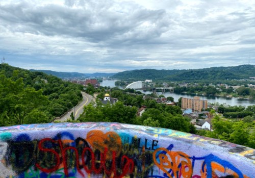 A scenic view of a river and hills, with colorful graffiti in the foreground and overcast skies above.