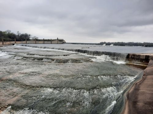 A serene view of a river with a low dam, featuring gentle waterfalls under a cloudy sky.