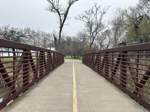 A metal bridge with a pathway leads through a park, surrounded by trees and a cloudy sky.