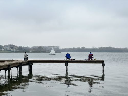 Three people sit on a dock by a calm lake, with a sailboat in the distance under a cloudy sky.