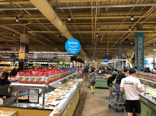 A busy grocery store aisle with shoppers, food displays, and a sign for a Prime member deal overhead.