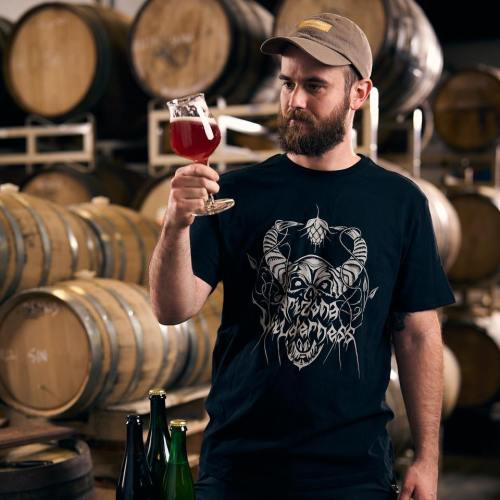A man in a black t-shirt examines a glass of red beer, surrounded by wooden barrels in a brewery.