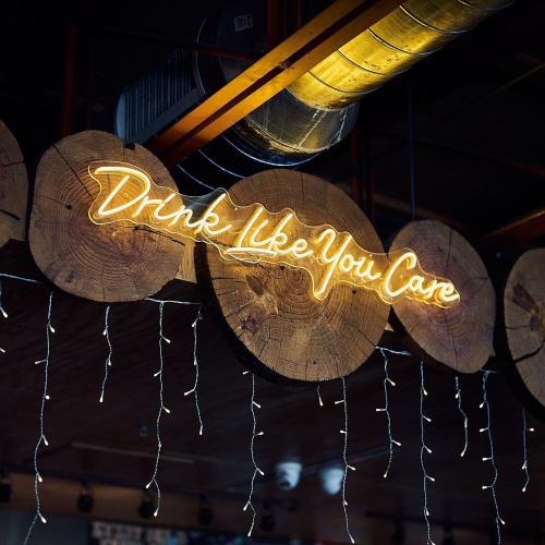 Neon sign on wooden circles reads "Drink Like You Care," with string lights hanging below.