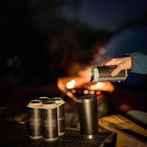 A hand pours a dark beverage from a can into a stainless steel tumbler, with a campfire glowing in the background.