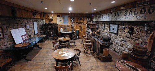 A rustic saloon interior with wooden tables, chairs, a bar, and stone walls, evoking an Old West atmosphere.