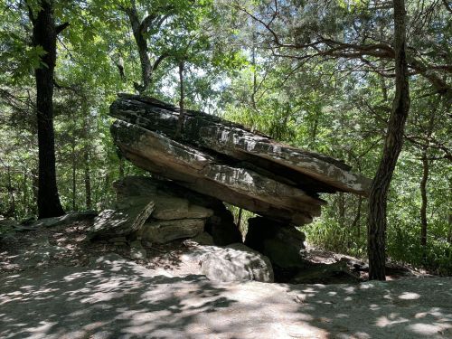 A large, balanced rock formation surrounded by trees and greenery in a natural setting.