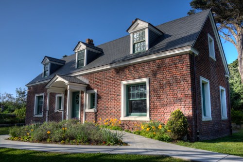 A charming brick house with a gabled roof, surrounded by a flower garden and a curved pathway.