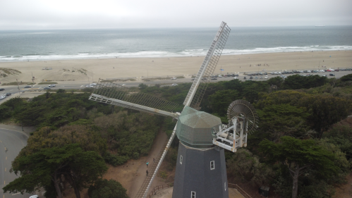 A windmill stands near the coast, overlooking a sandy beach and ocean under a cloudy sky.