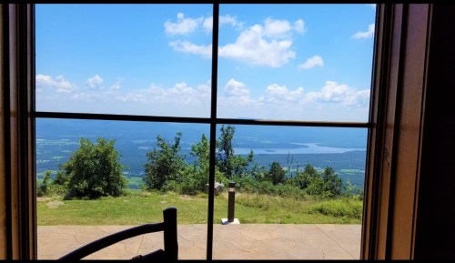 A scenic view of a lake and mountains through a large window, with blue skies and fluffy clouds above.