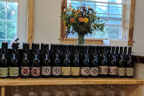 A row of beer bottles with decorative labels on a wooden shelf, alongside a vase of colorful flowers.