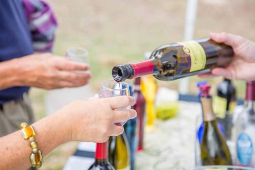 A hand pours red wine from a bottle into a glass at a tasting event, surrounded by various wine bottles.