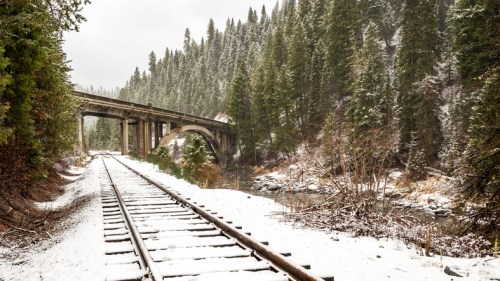 Snow-covered railway tracks lead through a forested area, with a bridge crossing over a river in the background.
