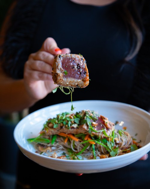 A person holds a piece of seared tuna above a bowl of colorful noodle salad with vegetables.