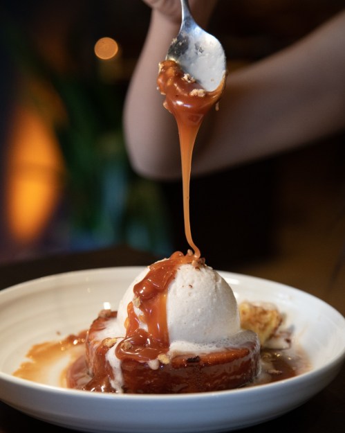 A spoon drizzles caramel sauce over a scoop of ice cream on a dessert plate.