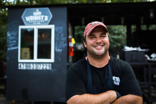 A smiling man in a cap stands with crossed arms in front of a food truck, surrounded by greenery.