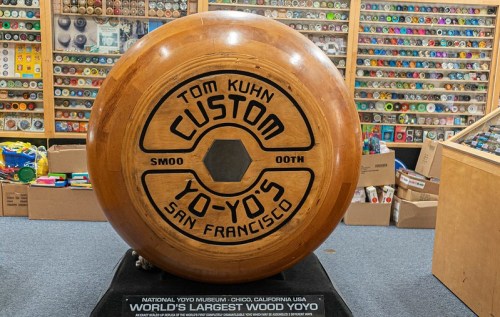 A large wooden yo-yo with "Tom Kuhn Custom Yo-Yo's San Francisco" engraved, displayed in a museum.