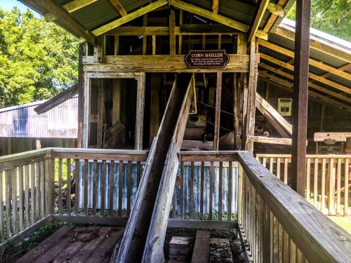 An old wooden corn sheller building with a sloped ramp and a sign, surrounded by greenery.