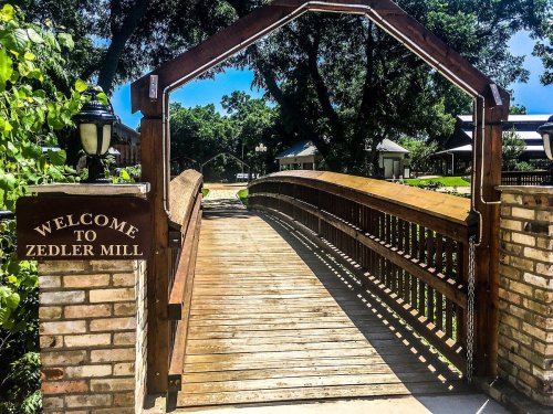 A wooden bridge with a sign reading "Welcome to Zedler Mill," surrounded by greenery and a clear blue sky.