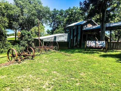 Rusty farm equipment in a grassy area near a weathered barn under a clear blue sky.
