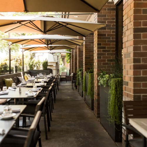 Outdoor dining area with tables set under large umbrellas, surrounded by greenery and brick walls.