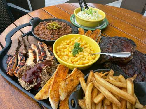 A platter of barbecue meats, macaroni and cheese, baked beans, coleslaw, and fries on a wooden table.