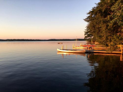 Serene lake at sunset with canoes and boats docked along the shore, surrounded by trees and calm water.