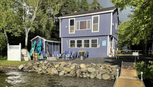 A blue two-story house by a lake, with a small shed and chairs on a rocky shore under a cloudy sky.
