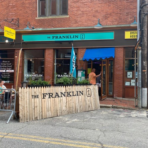 A brick building with a sign reading "The Franklin," featuring a blue awning and a wooden fence in front.