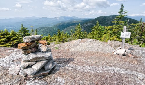 A rocky summit with a stacked stone cairn and a sign, overlooking a lush green mountain landscape under a cloudy sky.