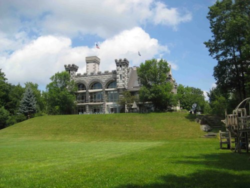 A large stone castle with turrets sits atop a grassy hill, surrounded by trees and under a partly cloudy sky.
