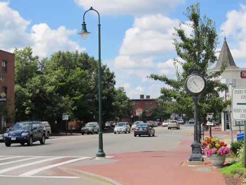 A street view of a small town with a clock, trees, and cars on a sunny day.